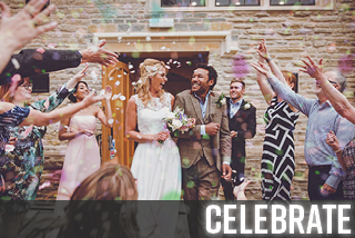A bride and groom walk out of the Tudor Hall door into the courtyard as colourful confetti is thrown all around them by delighted guests